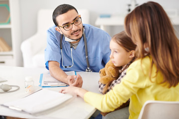 Portrait of young Middle-Eastern doctor talking to young mother trying to help her and her daughter during consultation in modern clinic