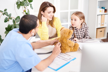 Back view portrait of young Middle-Easter doctor putting stethoscope to plush Teddy Bear toy during consultation with little girl and her mother in modern clinic.