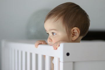 Close-up portrait of cute thoughtful baby staying in his baby cot holding the side and daydreaming,...
