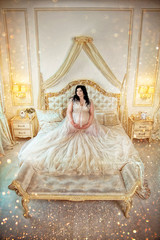 Charming pregnant brunette girl in long dress and diadem, sit on bed in luxurious bedroom