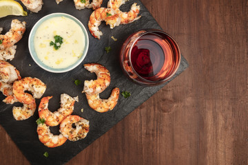 Overhead photo of plate of cooked shrimps on a dark rustic background, with a sauce and a glass of wine, with copy space