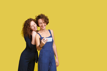 Pleased caucasian ginger girl gently smiling while her European friend making selfie. Indoor portrait of glad sisters with smartphone taking picture of herself on yellow background. Copy space