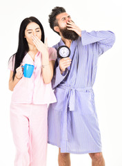 Couple in love, young family in pajama, bathrobe stand isolated on white background. Girl with coffee cup, man hold clock in hand and yawning. Perfect morning concept. Couple woke up early in morning.