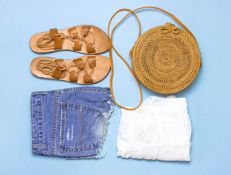 Fashionable handmade natural organic rattan bag, leather sandals, chamomile flowers on blue background flat lay. Copy space, top view. Ecobags from Bali. Eco-bag concept.