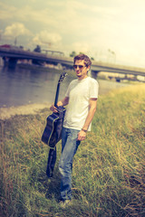 Naklejka na ściany i meble Junger Mann mit Sonnenbrille steht an Flussufer, Gitarre in der Hand