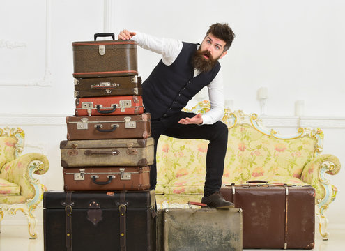 Man, butler with beard and mustache delivers luggage, luxury white interior background. Macho elegant on shocked face standing near pile of vintage suitcase. Luggage and travelling concept.