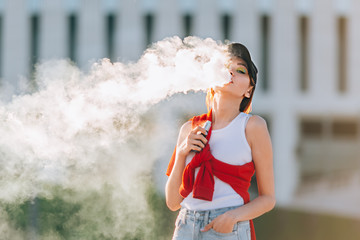 Pretty young hipster asian woman in black hat vape ecig, vaping device at the sunset. Toned image.
