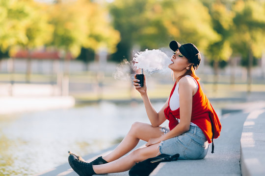 Pretty young hipster asian woman in black hat vape ecig, vaping device at the sunset. Toned image.