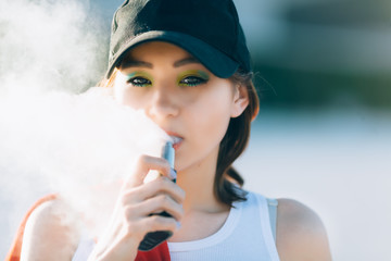 Pretty young hipster asian woman in black hat vape ecig, vaping device at the sunset. Toned image....