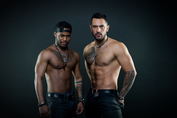 Fototapeta na wymiar Machos with muscular tattooed torsos look attractive, dark background. Masculinity concept. Athletes on confident faces with nude muscular chests. Guys sportsmen with sexy muscular torsos