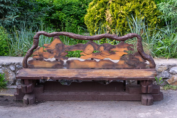 A wooden bench made with hand made of lumber and boards painted with brown paint with a carved pattern of animals and birds on the back against the background of green trees and bushes