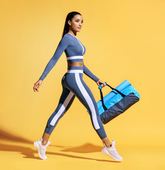 Fototapeta na wymiar Go to training. Sporty woman with bag on yellow background. Dynamic movement. Side view. Sports and healthy lifestyle