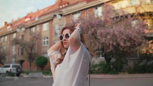 Young beautiful fashionable woman wearing stylish round sunglasses, white chiffon blouse, wrist watch walking in street