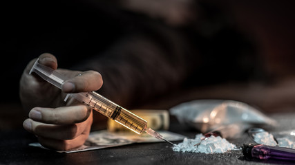 Male junkie hand holding drug injection syringe while lying near heroin powder, spoon and cigarette lighter for heroin cooking and money on dark floor. Hard drug addiction concept - obrazy, fototapety, plakaty
