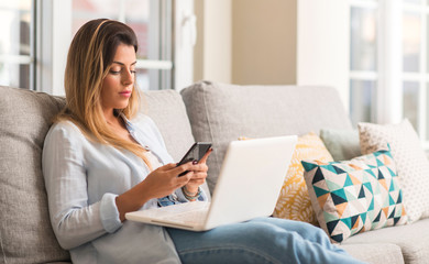 Fototapeta premium Young beautiful woman smiling using laptop and phone on the sofa relaxed.