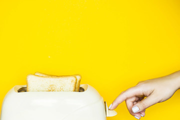 Toasted bread sticks out of the toaster on a yellow background. Female hand switch on the toaster