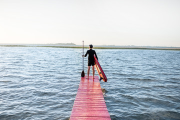 Landscape view on the lake with man standing with paddleboard on the pier during the sunrise