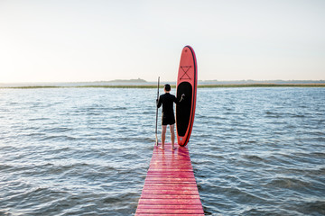 Landscape view on the lake with man standing with paddleboard on the pier during the sunrise