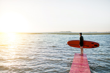 Landscape view on the lake with man standing with paddleboard on the pier during the sunrise
