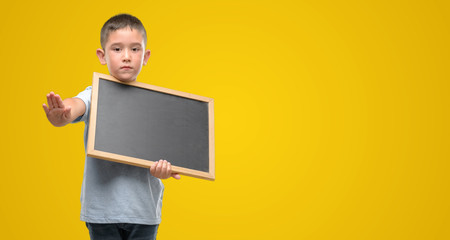 Dark haired little child holding a blackboard with open hand doing stop sign with serious and confident expression, defense gesture