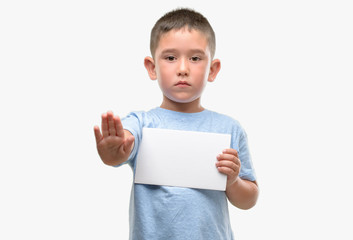 Dark haired little child holding a blank card with open hand doing stop sign with serious and...