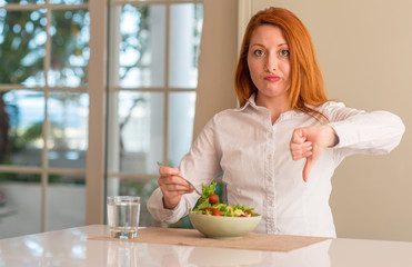 Redhead woman eating fresh green salad at home with angry face, negative sign showing dislike with thumbs down, rejection concept