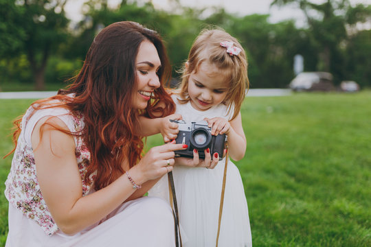 Smiling woman in light dress and little cute child baby girl holding retro vintage photo camera in green park. Mother, little kid daughter. Mother's Day, love family, parenthood, childhood concept.