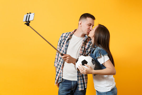 Kiss couple, woman man, football fans doing selfie on mobile phone with monopod selfish stick, cheer up support team, soccer ball isolated on yellow background. Sport family leisure lifestyle concept