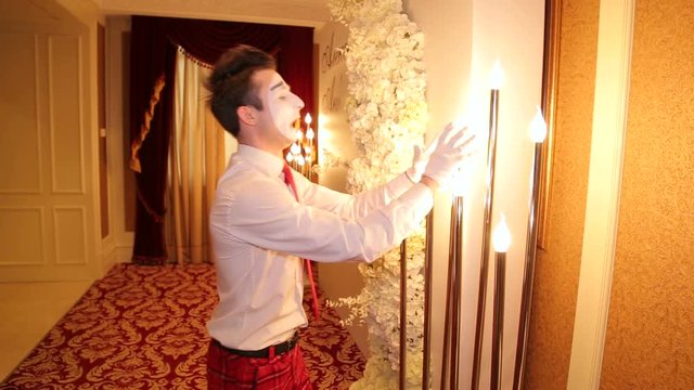 Portrait of a mime man with a white face on the holiday, he pretends to warm his hands near small lamps. Mime artist with a white suit is working on the holiday.