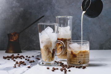 Iced coffee cocktail or frappe with ice cubes and pouring cream in three different glasses with vintage jezva and coffee beans around on white marble table with grey concrete wall at background.