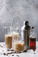 Iced coffee cocktail or frappe with ice cubes and cream in different glasses with silver shaker, bottle of rum, coffee beans around on white marble table with grey concrete wall at background.