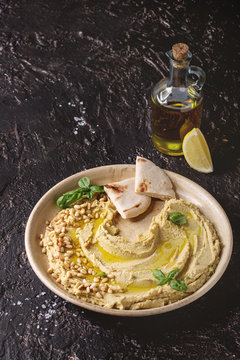 Homemade traditional spread hummus with pine nuts, olive oil, basil served on ceramic plate with pita bread and olive opl over dark texture background. Mediterranean snack.