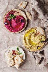 Variety of homemade traditional and beetroot spread hummus with pine nuts, olive oil, pomegranate served on ceramic plates with pita bread and green salad on linen cloth background. Flat lay, space.
