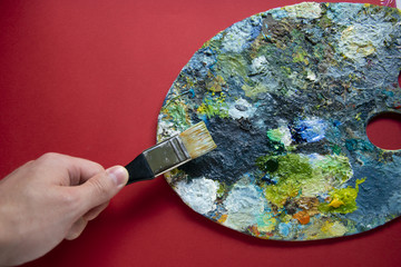 Artist's hand with a brush and a palette of oil paints on a red background