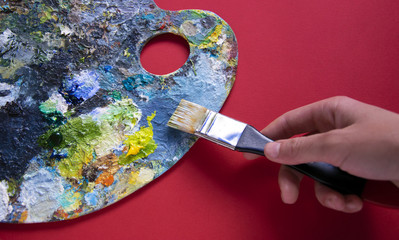 Artist's hand with a brush and a palette of oil paints on a red background