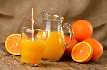 Fototapeta na wymiar Glass with orange juice and straw, jug with fresh juice and pile of oranges in the background on wooden table