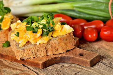Close-up of wholegrain slices of bread with scrambled eggs, fresh herbs, spring onions and tomatoes in background