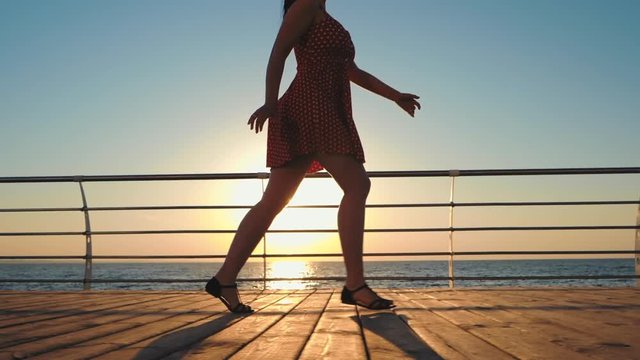 Young woman in retro short dress running on wooden embankment near sea. Girl walking at sunrise or sunset background. Beautiful scene of female legs. Slow motion.