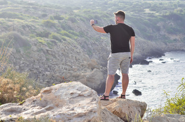 Man making photo with his mobile phone camera standing on top of cliff in summer mountains. Cyprus.