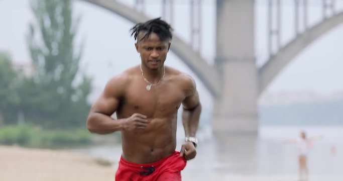 Sexy macho man with muscular body running on beach under rain