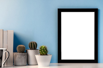 Blank black frame with cactuses, books and a black mug with coffee on the background of a bright blue wall