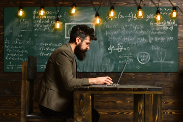Businessman in suit with laptop at school desk. Bearded man work on laptop in classroom. Man with long beard with notebook on chalkboard. Scientist in glasses on genius face. New technology concept