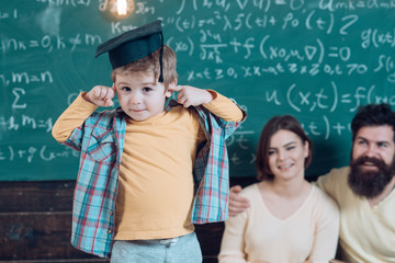 Wunderkind concept. Smart child, wunderkind in graduate cap pointing at his head. Parents listening their son, chalkboard on background. Boy presenting his knowledge to mom and dad