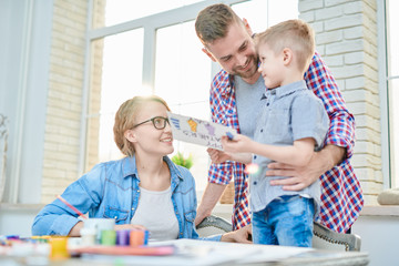 Portrait of modern young family with cute little son smiling happily looking at each other while  celebrating Fathers day together  standing against big windows at home