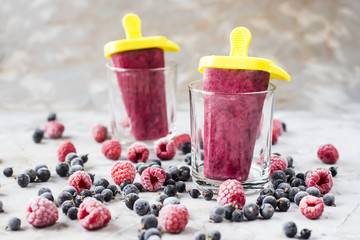 Berry ice cream on a stick in transparent glasses. Homemade of raspberries and currants, cooked with special forms. On a gray table among frozen berries