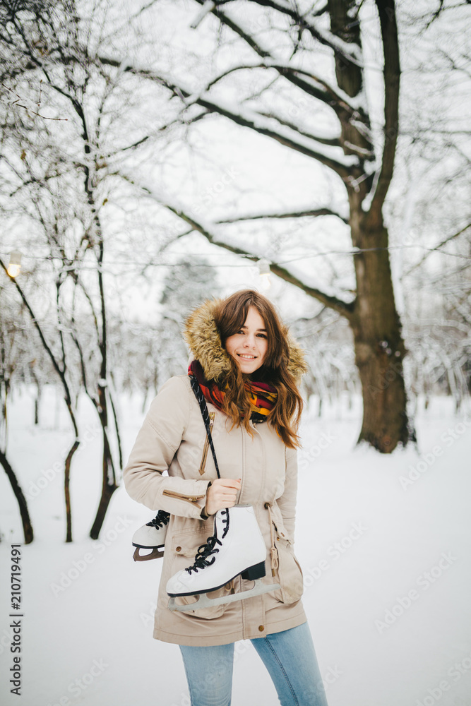 Wall mural Theme is weekend holiday in winter. A beautiful young Caucasian woman stands in a snow covered park in jacket with hood and fur in jeans and holds on shoulder pair of white skates for long shoelaces