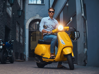 Fototapeta na wymiar Young stylish guy dressed in a in a white shirt and jeans ride on yellow classic italian scooter on an old Europe street in the evening.