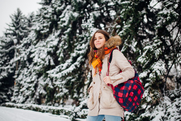 Theme is weekend holiday in winter. A beautiful young Caucasian woman stands in snow park in jacket with hood and fur in jeans and smiles with checkered backpack on background of New Year tree