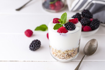 White yogurt with muesli and raspberries in glass bowl on white wooden background.