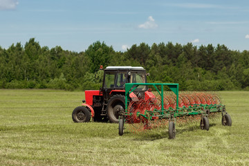 Agricultural machinery, a tractor collecting grass in a field against a blue sky. Hay harvesting, grass harvesting. Season harvesting, grass, agricultural land.
